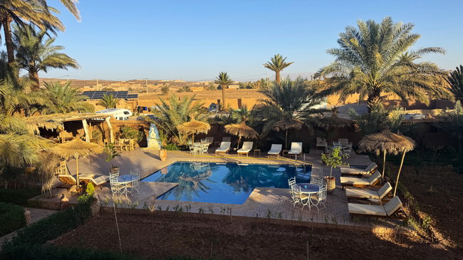 Voyage moto au Maroc MM8 – Petit-déjeuner dans le jardin verdoyant de l'hébergement Espace petit-déjeuner de l'hébergement avec coin salon couvert et jardin verdoyant dans le paysage aride du sud du Maroc