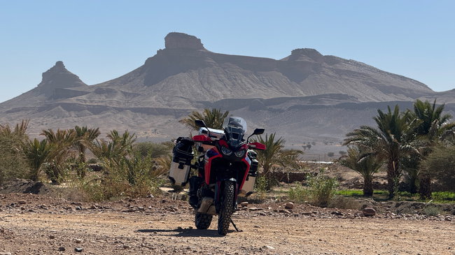 Voyage moto au Maroc MM8 – À travers le vaste désert de pierres du sud du Maroc Vaste désert de pierres au sol aride, avec quelques montagnes à l'horizon et une route isolée